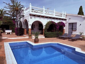 A pool with a white balustrade, a covered patio area, and a building with arches and a balcony.