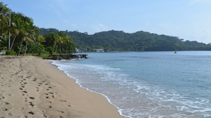 Ein Strand mit Fußspuren, die zum Wasser führen, Palmen und eine bewaldete Hügelkette im Hintergrund.
