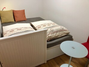 A small room with a bed, a white radiator, a small round table, and a red chair.