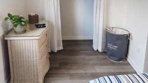 A small, neatly organized room with a wicker table, a potted plant, a television mounted on the wall, a closet with hanging clothes, and a laundry basket.