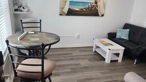 A living room with a round table, a black sofa, a white coffee table, and a painting of a beach scene.