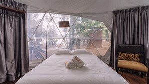 A bed with white linens, a pair of towels, and a chair with a patterned cushion in a room with a large window and curtains. - Roxy Telok Serabang (Lundu)