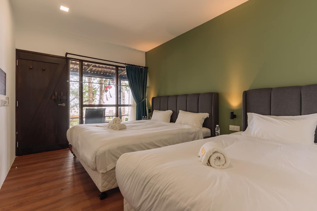 A hotel room with two beds, a wooden floor, a green wall, and a window with curtains.