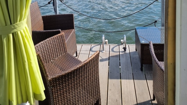 Een scheepdek met rieten meubilair, een uitzicht op een meer en een beboste oever.