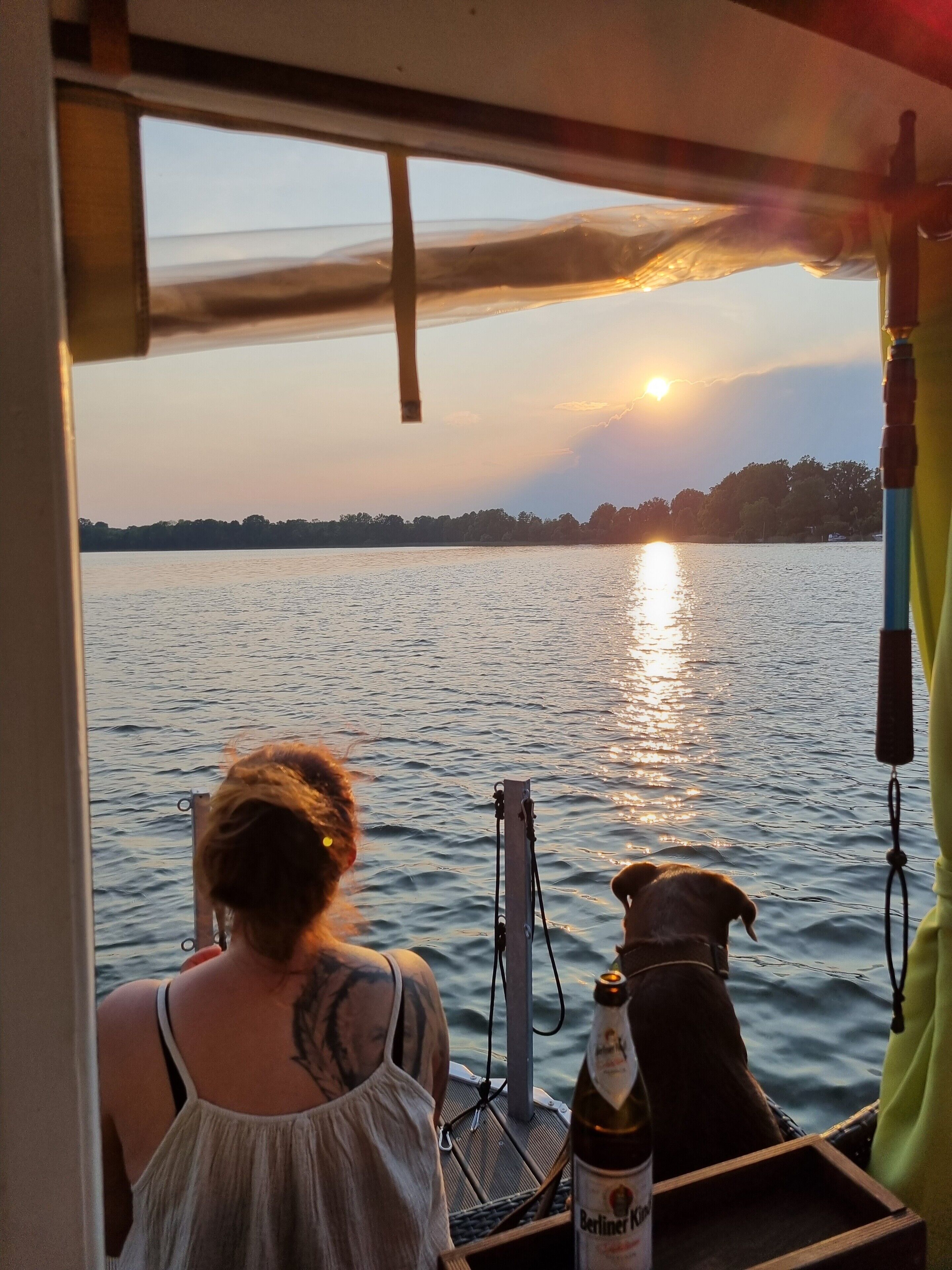 A boat with a person and a dog, a bottle of beer, and a view of the sunset over water.