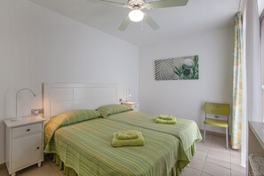 Une chambre avec deux lits, une table de chevet, une chaise, un ventilateur de plafond et une œuvre d’art murale.
