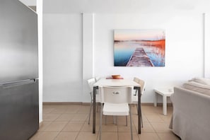 A modern kitchen with a refrigerator, a dining table with chairs, and a painting of a sunset over water.