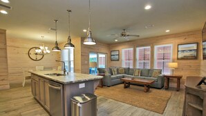 A modern kitchen with a central island, a dining area with a sofa and coffee table, and a living room with a couch and a ceiling fan. - Hammock Dunes J (Orange Beach)
