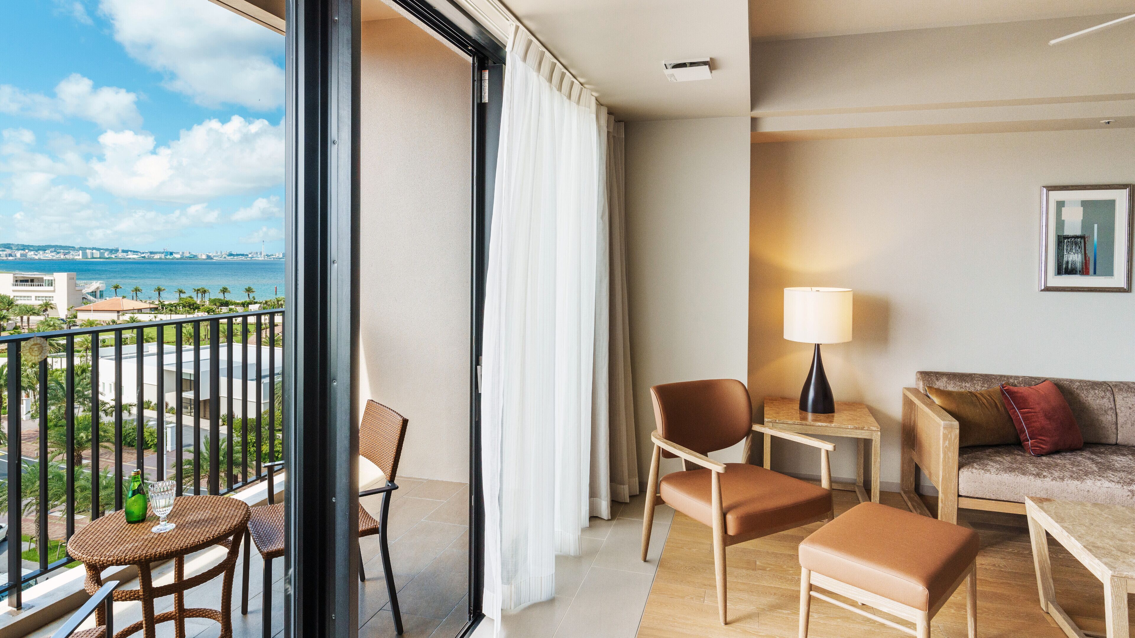 Un balcon offrant une vue sur la mer et la ville, aménagé avec des fauteuils en osier, un canapé et une petite table avec une lampe.