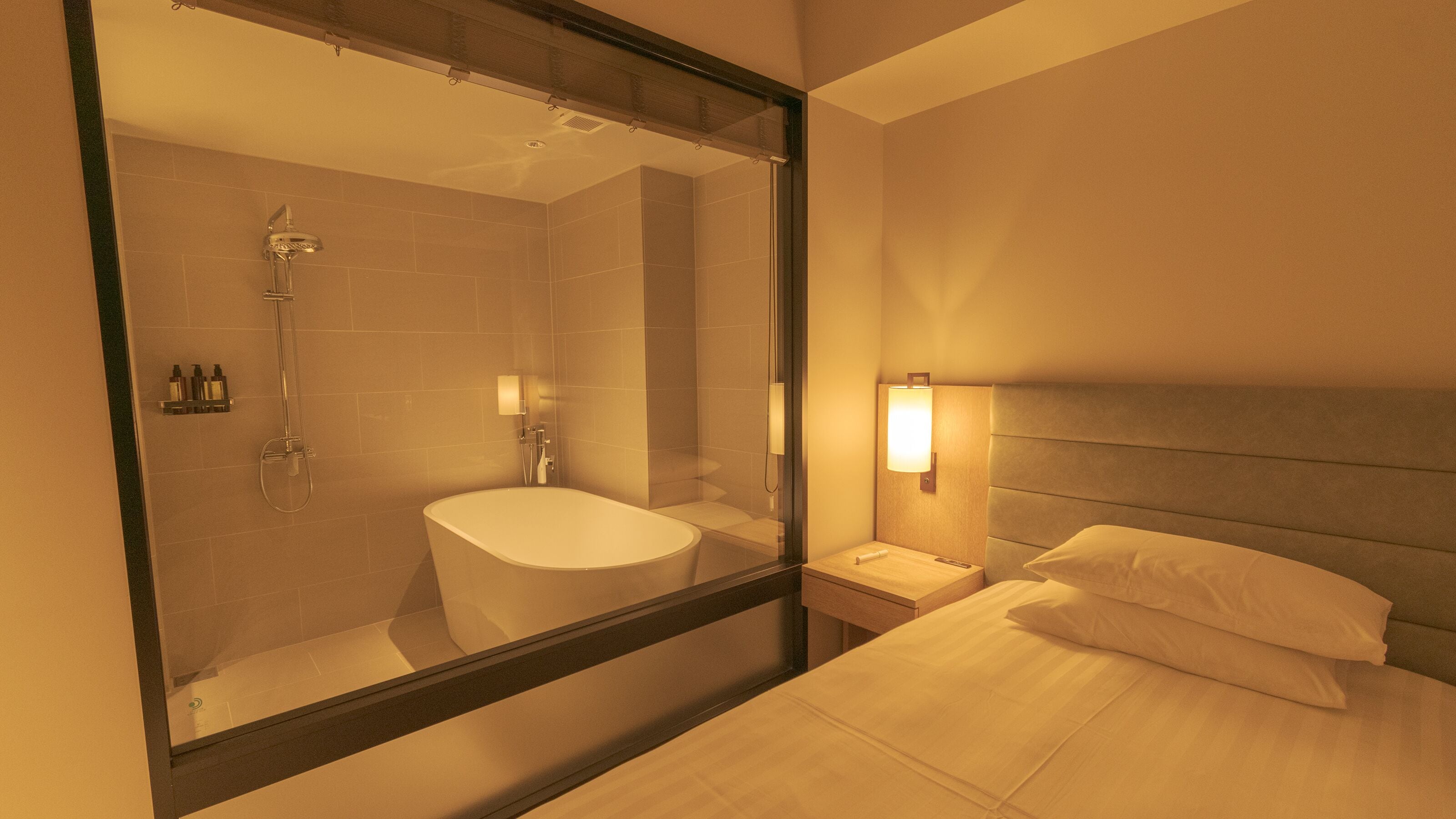 Une chambre d’hôtel avec un grand miroir, une baignoire, une table de chevet avec une lampe et un lit avec des oreillers.