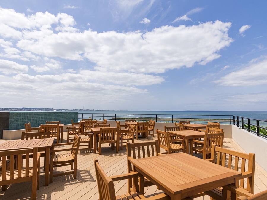 A terrace with wooden tables and chairs, overlooking a coastal view.