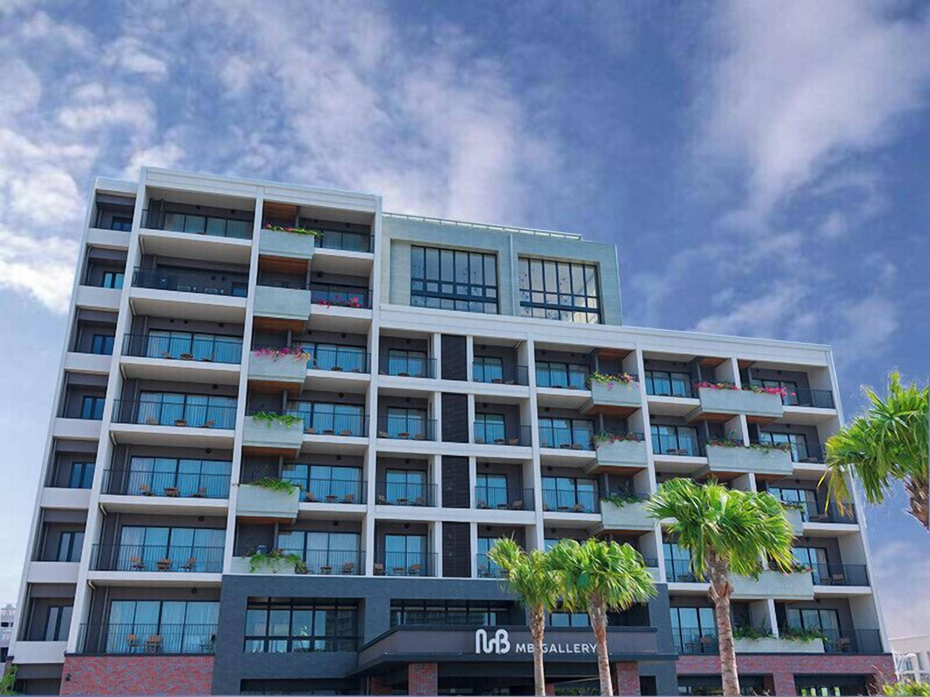 A multi-story hotel with balconies, a storefront labeled "MB GALLERY," and palm trees in the foreground.