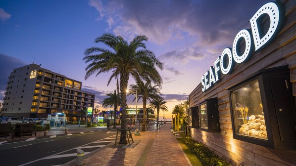 A coastal street with a seafood restaurant, a multi-story hotel, and palm trees. - MB GALLERY CHATAN by THE TERRACE HOTELS (Chatan)