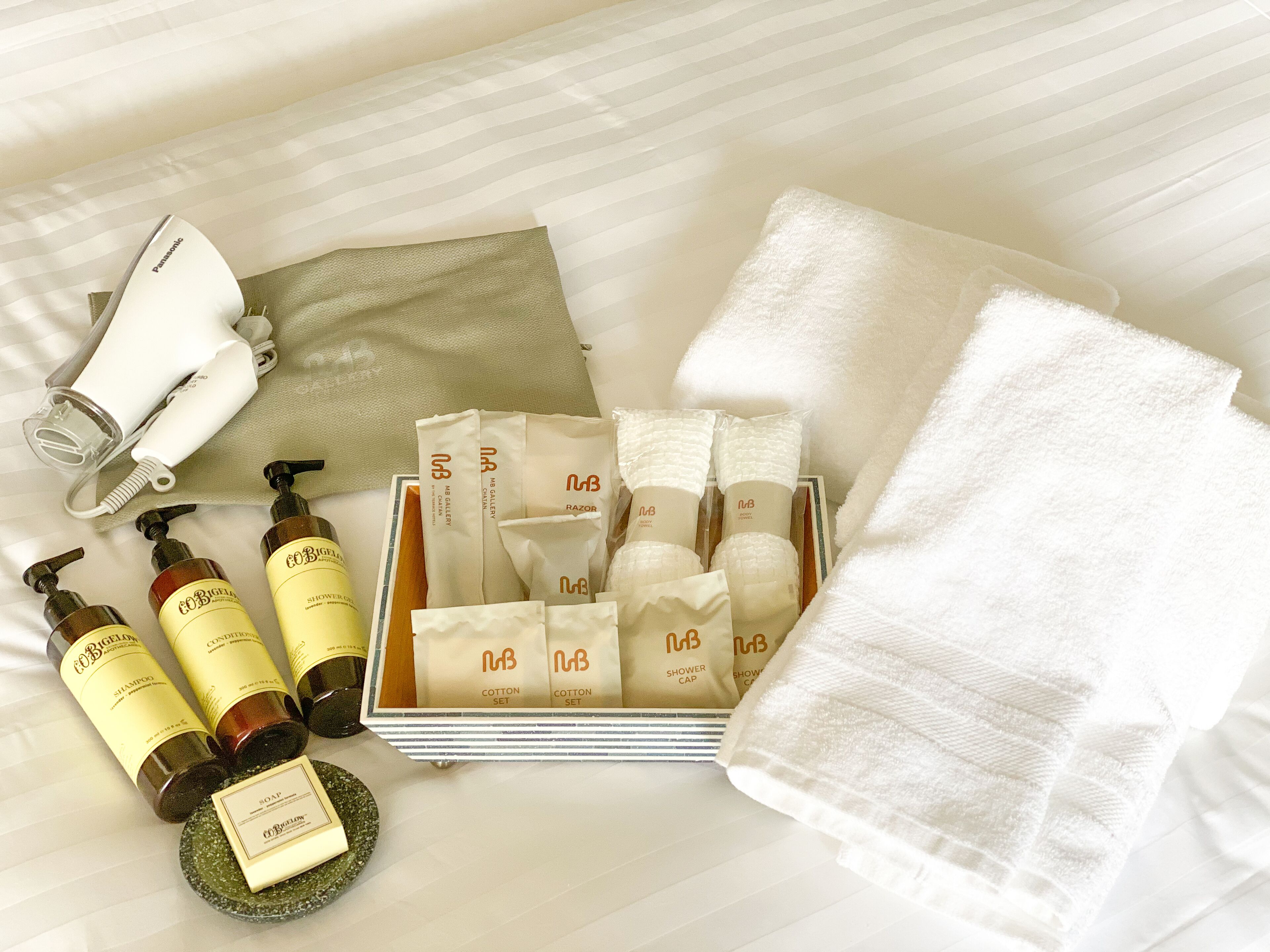 Un conjunto ordenado de artículos de aseo, incluyendo champú, acondicionador, gorro de ducha y toallitas de algodón, sobre una cama con una toalla blanca y un secador de pelo.
