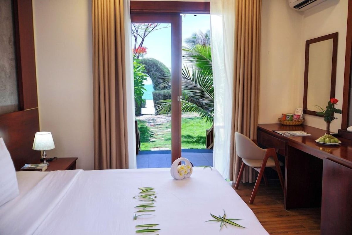 A hotel room with a bed, a desk, a chair, a lamp, a mirror, and a view of a garden and water.