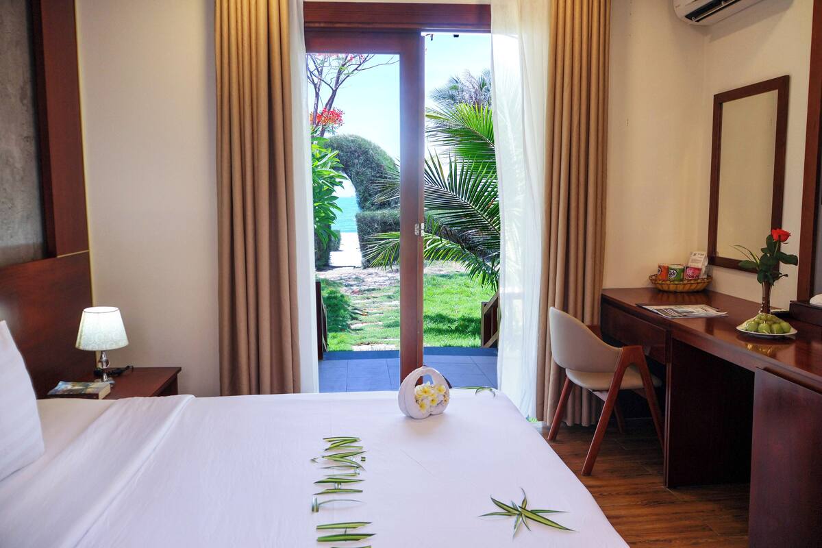 A hotel room with a bed, a desk, a chair, and a view of a garden and ocean.
