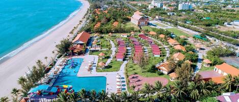 An aerial view of a resort with a large swimming pool, beach, and palm trees.