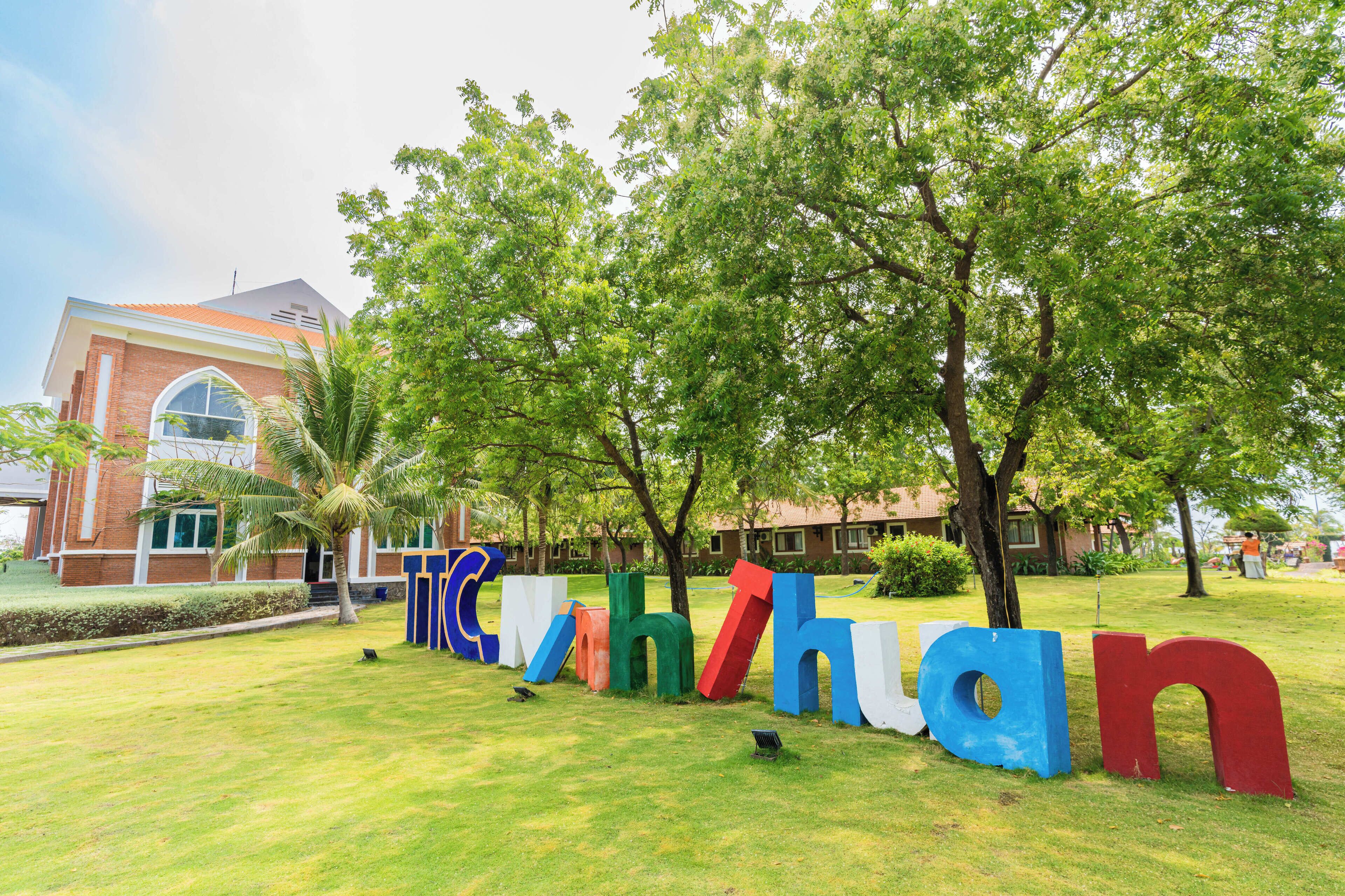 A grassy area with colorful letters spelling "TRINITY" and a building with a red roof in the background.