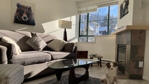 A living room with a sofa, a glass coffee table, a dog, a fireplace, and a window with a view of trees. - Cozy, Bright Whistler Village Studio, Hot Tub, Sleeps 4 ( 2 adults 2 children)  (Whistler)