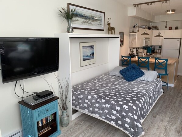 Une chambre à coucher de taille réduite, avec un lit, une télévision, un petit meuble et, au fond, un coin cuisine.