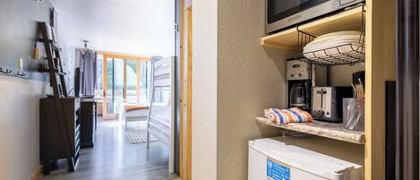 A kitchen with a microwave, toaster, and refrigerator. There is a wooden shelf above the counter and a door leading to a balcony.