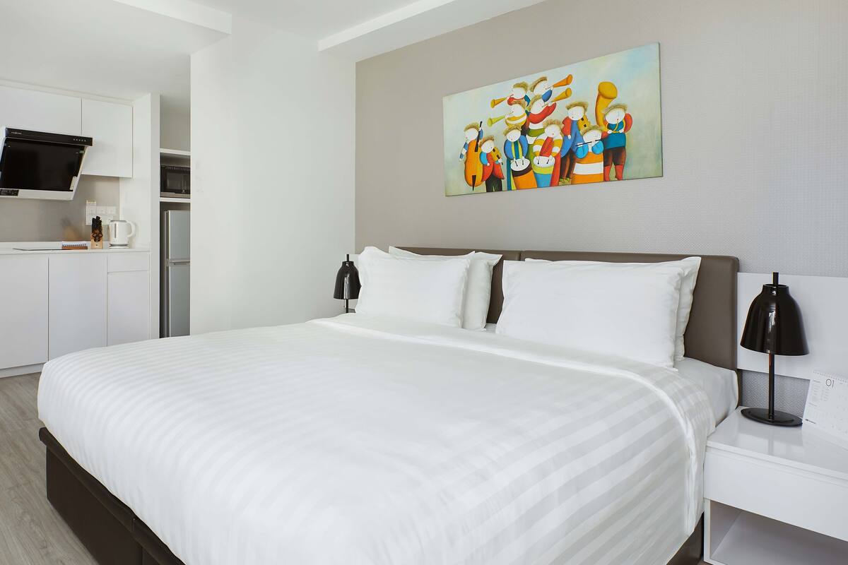 Quarto de hotel com uma cama grande, mesa de cabeceira, escrivaninha, cadeira, máquina de lavar, micro-ondas e um quadro na parede.