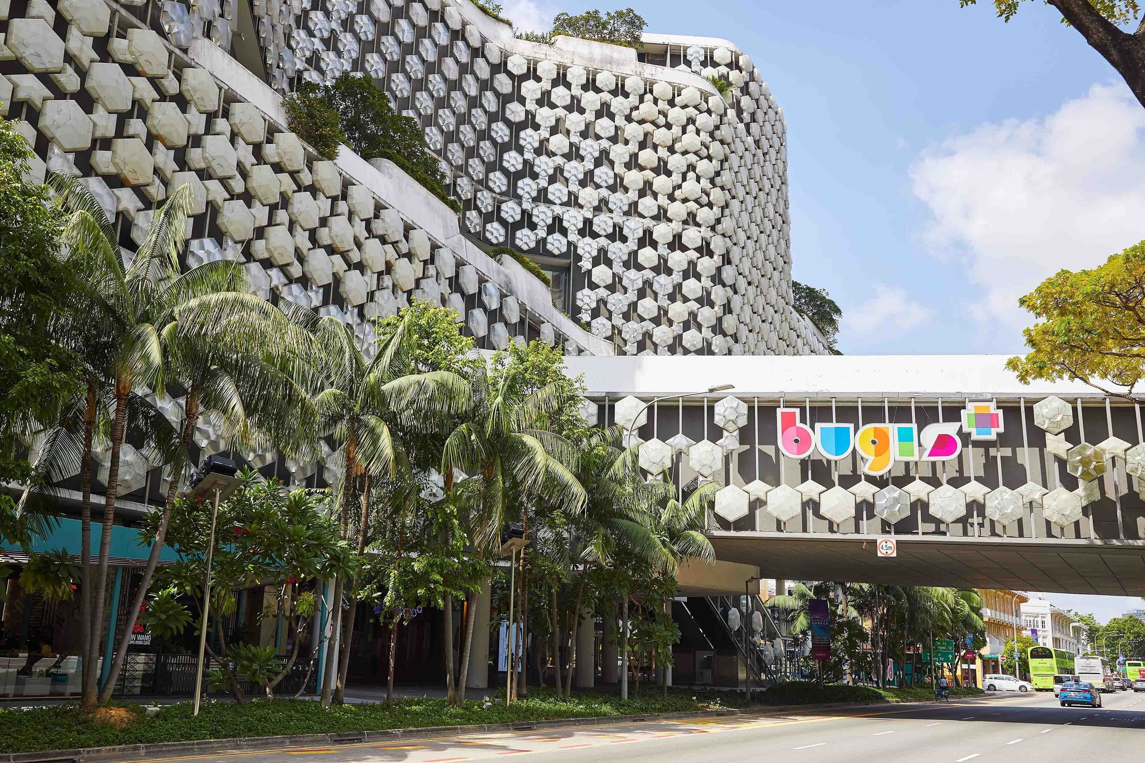 A modern building with a unique hexagonal facade and a pedestrian overpass with the word "bugis" on it.