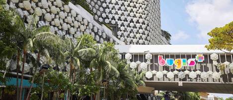A modern building with a unique hexagonal facade and a pedestrian overpass with the word "bugis" on it.