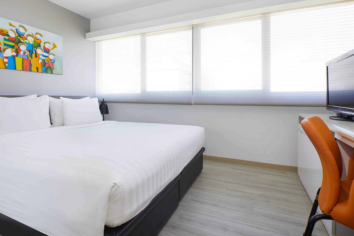 Quarto de hotel com uma cama grande, uma mesa com computador, televisão e um quadro colorido na parede.