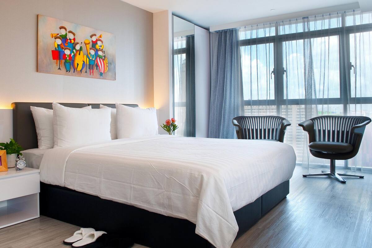 Quarto de hotel moderno com uma cama grande, mesa de cabeceira com relógio, uma cadeira e um quadro na parede.