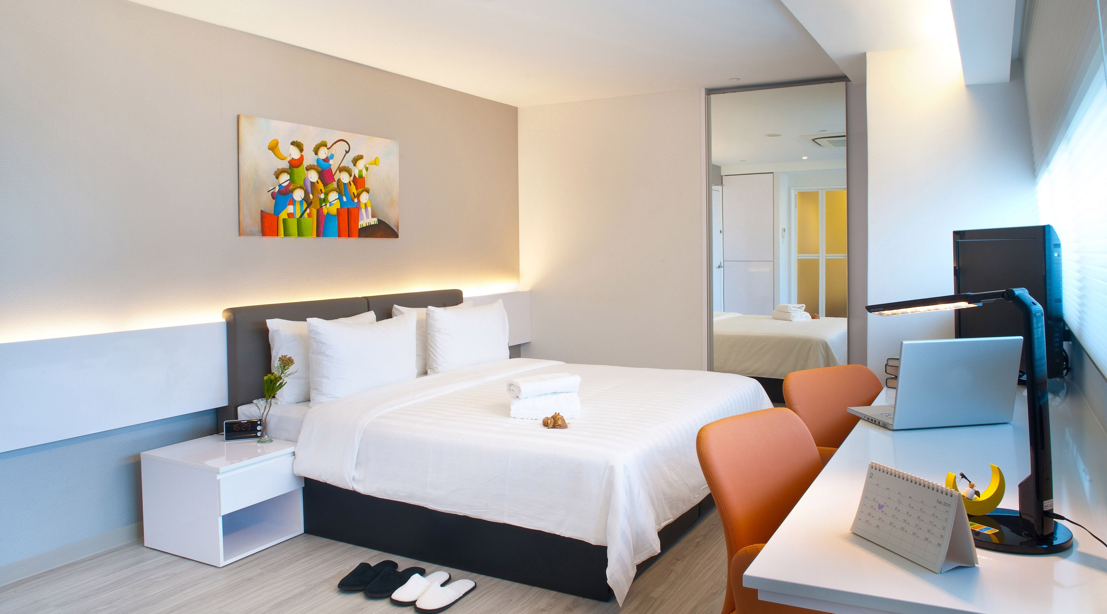 A modern hotel room with a large bed, a desk with a laptop, a chair, a nightstand, and a wall-mounted painting.