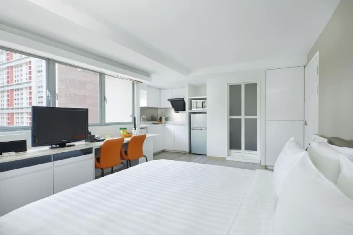 A hotel room with a bed, a desk with a TV, a small kitchen area, and a window with a city view.