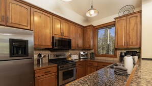 Cocina con armarios de madera, electrodomésticos de acero inoxidable, encimeras de granito y ventana con vistas a árboles.