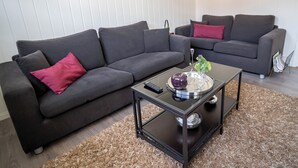 A living room with two dark grey sofas, one with a red pillow, a glass coffee table with a remote control, and a decorative bowl.