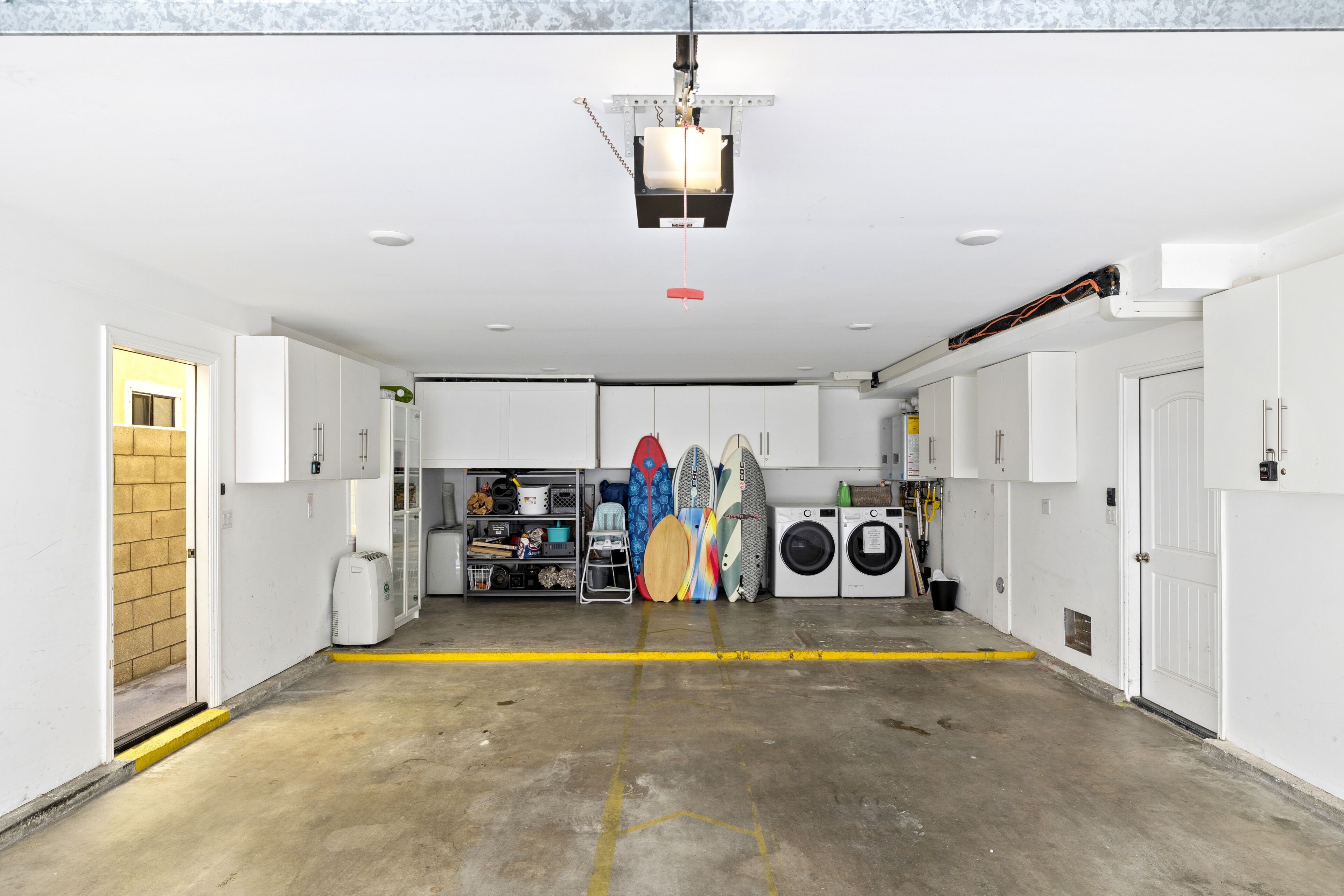 Un espace intérieur propre et bien éclairé, avec des murs blancs, un sol en béton et un luminaire fixé au plafond. On y trouve des planches de surf appuyées contre le mur, un lave-linge et un petit coin salon avec une chaise et une table.