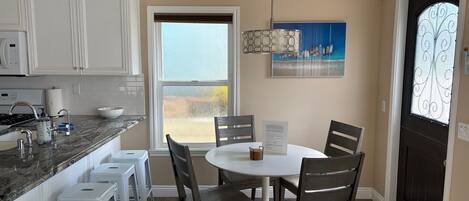 A kitchen with white cabinets, a marble countertop, a dining table with chairs, a window with a view, and a door with a decorative glass panel.