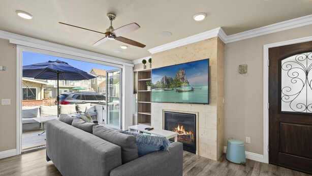 A living room with a grey sofa, a fireplace, a ceiling fan, and a large window overlooking a backyard.