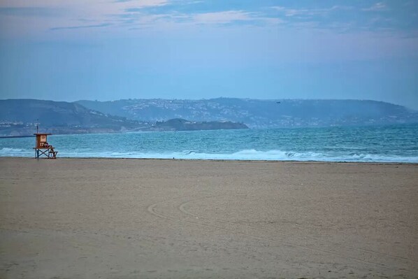 Una playa con una torre de salvavidas y una silla, cielo despejado y un paisaje montañoso distante.