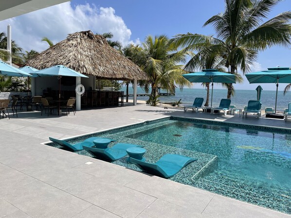 A pool area with lounge chairs and umbrellas, a bar area with a thatched roof, and a view of the ocean.