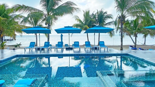 A pool area with blue chairs and umbrellas, surrounded by palm trees and a view of the sea. - BLUE DIAMOND TRANQUILITY RESORT-Private Luxury Villa - 8 King Rooms-ON THE BEACH (Stann Creek District)