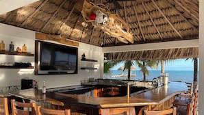 A bar area with a thatched roof, wooden chairs, a large TV, and a view of the ocean. - BLUE DIAMOND TRANQUILITY RESORT-Private Luxury Villa - 8 King Rooms-ON THE BEACH (Stann Creek District)
