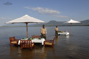 A dining setup on a boat with a table and chairs, surrounded by water with mountains in the background.