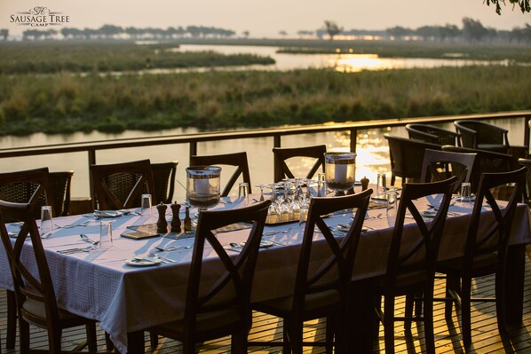 A dining table set for six with a view of a water body at sunset.