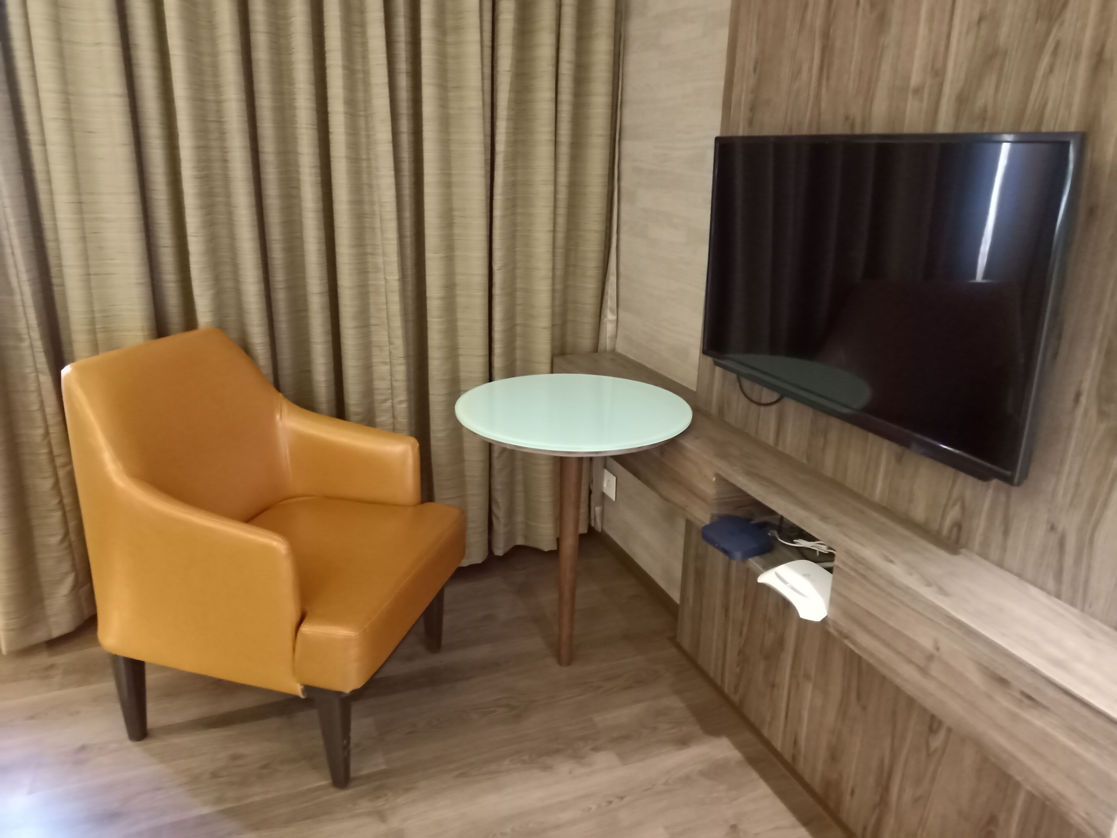 A room with a brown leather chair, a small round table, a flat-screen TV mounted on a wooden wall, and a power outlet with a blue device plugged in.