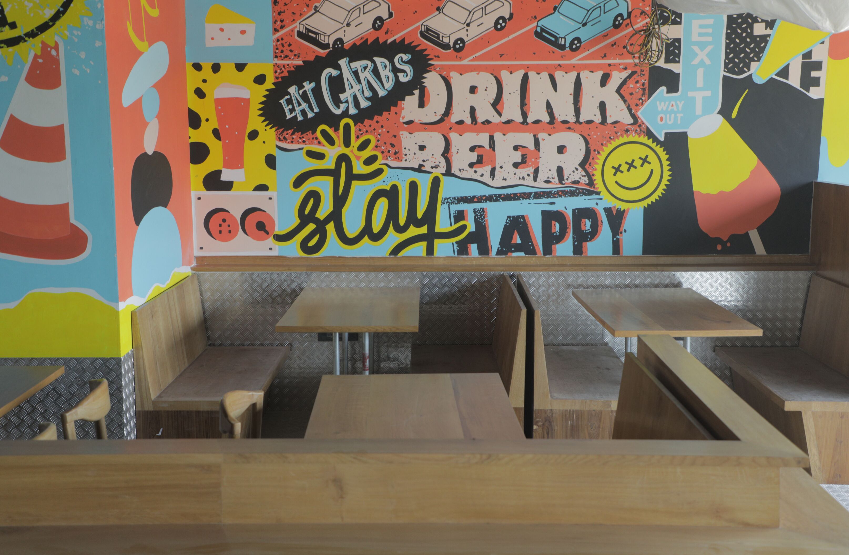 A restaurant interior with wooden tables and benches, colorful wall art, and a sign that reads "Eat Carbs Drink Beer Stay Happy".