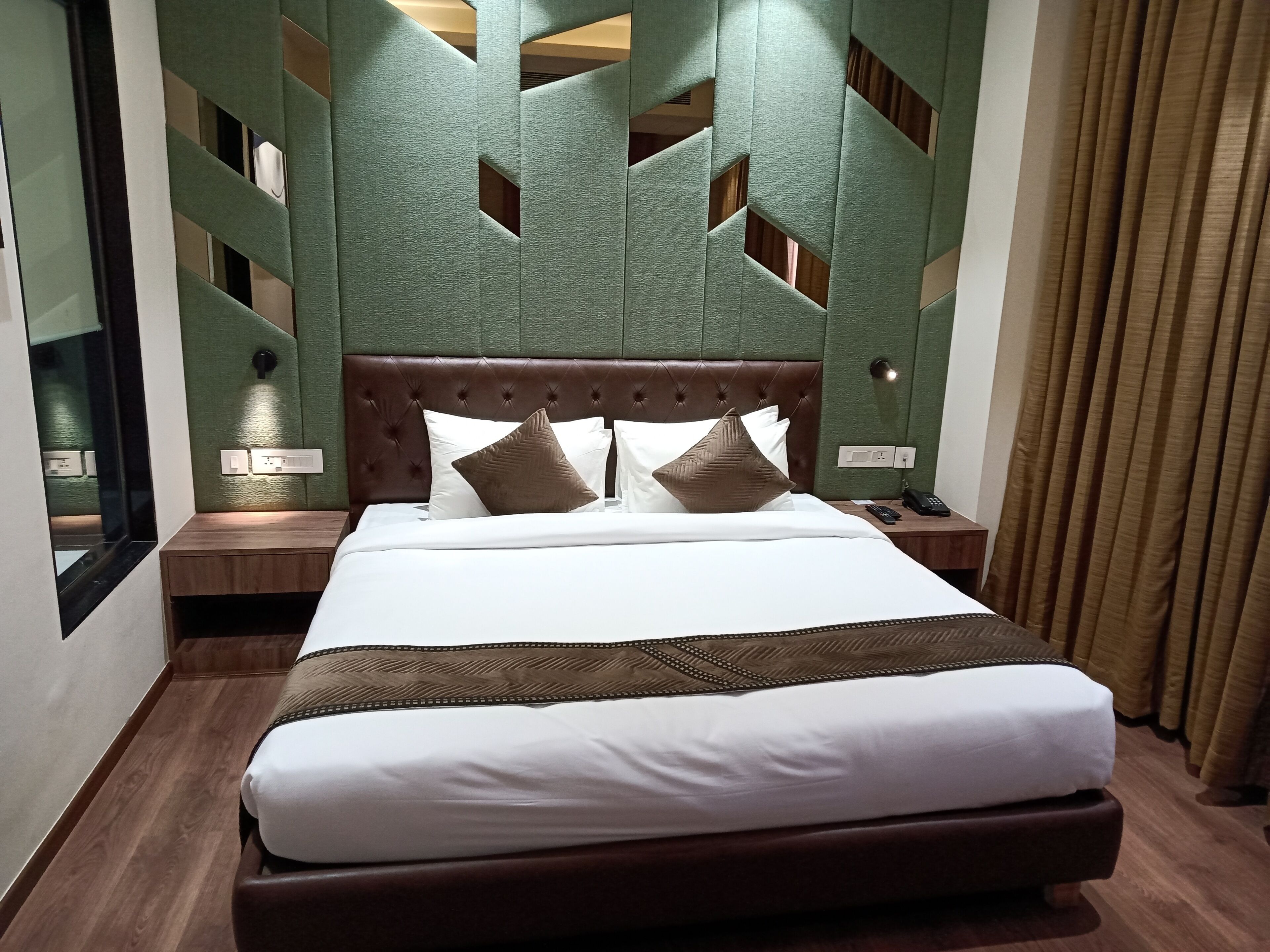 A neatly made bed with a brown headboard, white bedding, and decorative pillows. There is a nightstand with a phone and a lamp beside the bed. The room features a green textured wall with geometric cutouts and a brown curtain on the window.