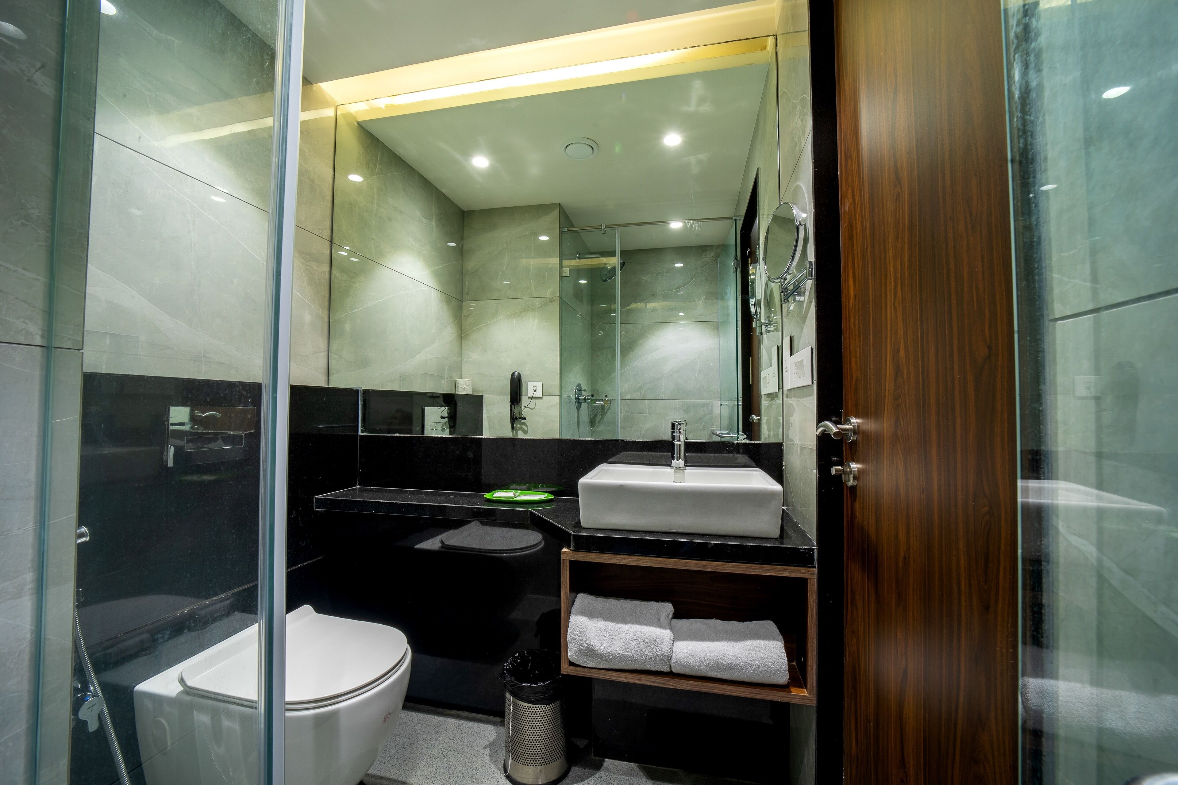 A modern bathroom with a glass-enclosed shower, a white toilet, a black sink, and a wooden vanity with a mirror.