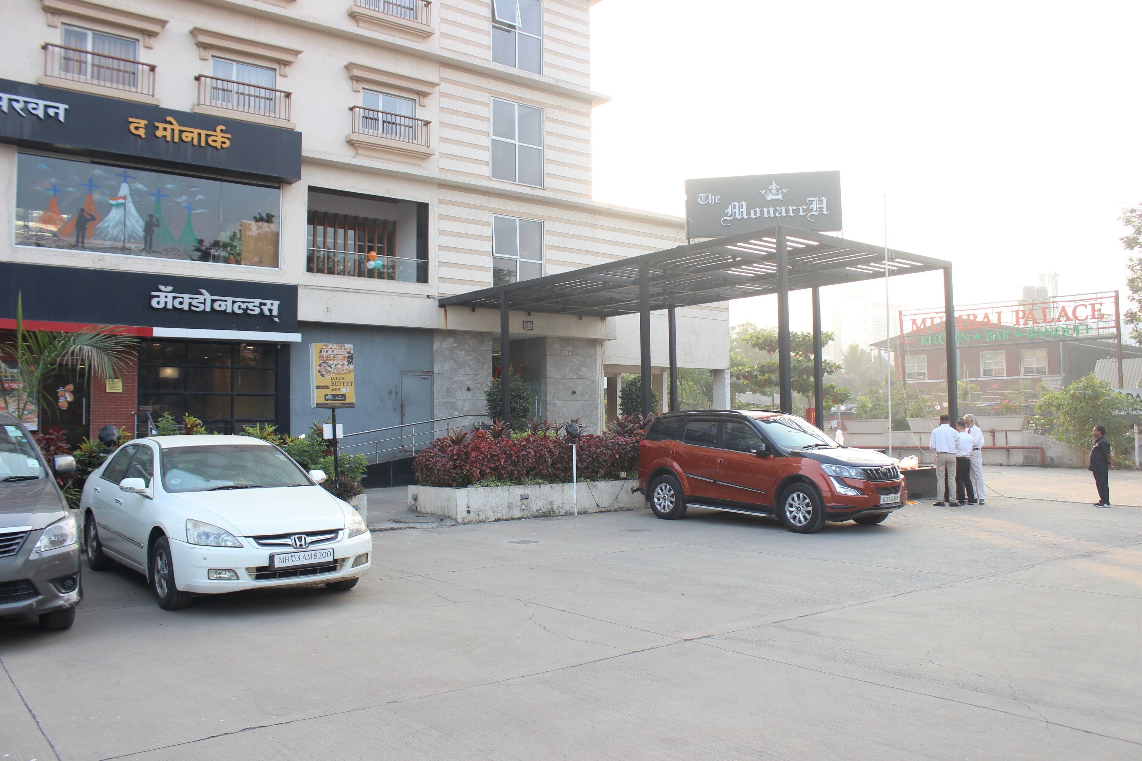 Entrada de un hotel con un corredor cubierto, un coche blanco y un coche rojo estacionados al frente.