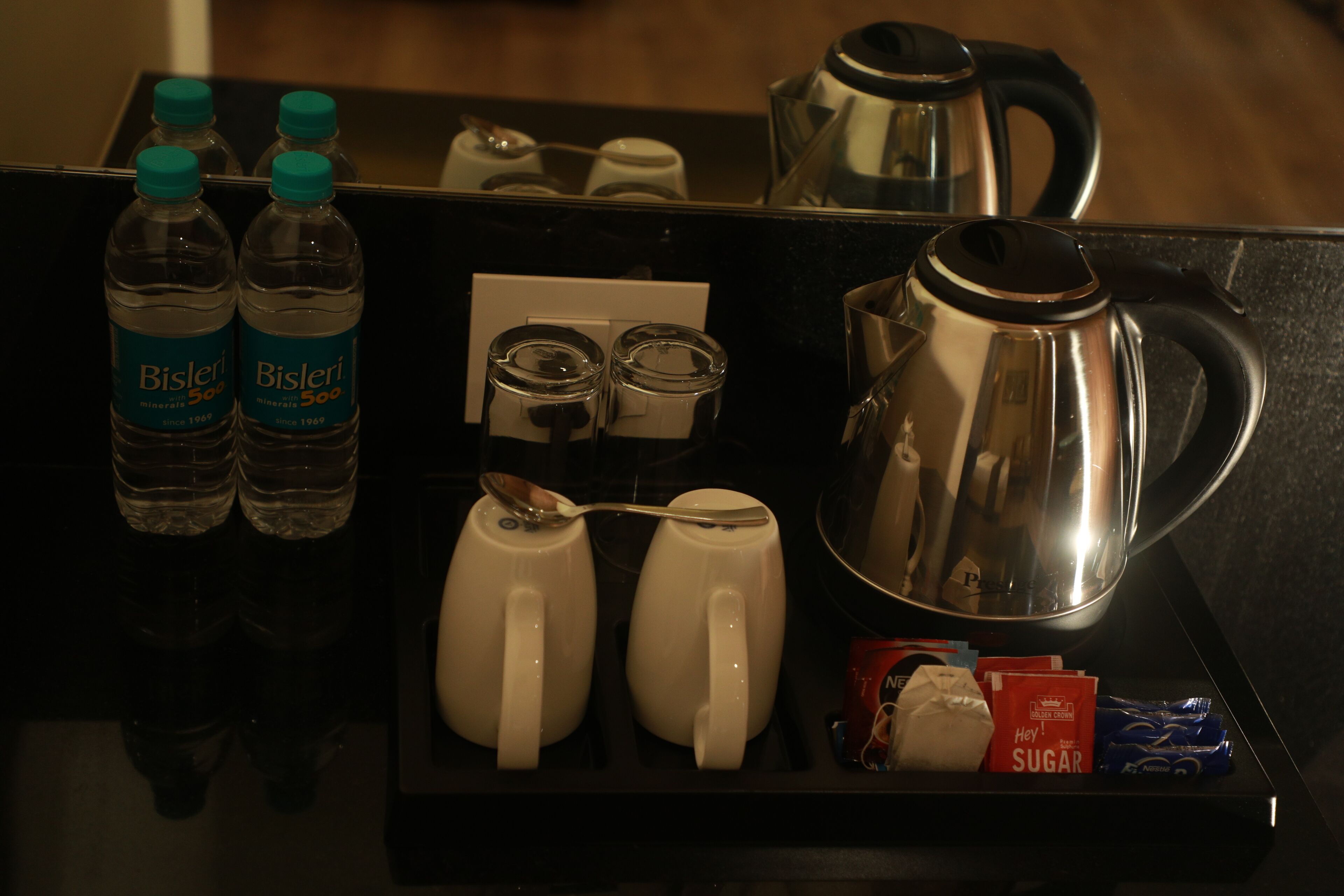 A tray with Bisleri mineral water bottles, two white mugs, a stainless steel kettle, sugar packets, and a spoon.