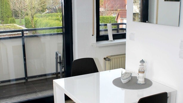 Ein moderner Essbereich mit einem weißen Tisch und schwarzen Stühlen, Blick ins Freie durch ein großes Fenster und einer herzförmigen Dekoration an der Wand.